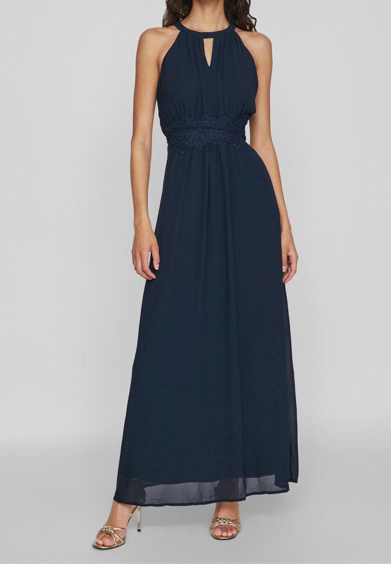 Femme portant une robe longue bleue marine sans manches à col halter avec une ceinture en dentelle et des talons à brides tressées dorées, debout sur un fond gris.