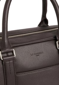 Bolso de mano de cuero marrón con una textura de grano, que cuenta con herrajes de plata, un cierre de cremallera y un logotipo de marca grabado en la parte frontal.