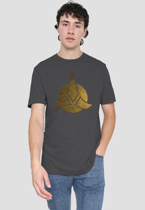 Dark grey cotton t-shirt featuring a metallic gold emblem, circular design with pointed accents, short sleeves, and a relaxed fit.