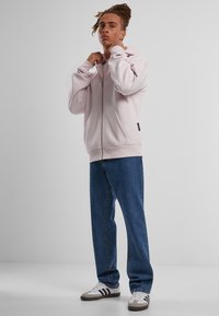 Felpa con zip rosa chiaro in morbido tessuto, dotata di un ampio cappuccio e tasche frontali, abbinata a jeans blu larghi e sneaker grigie con strisce nere.