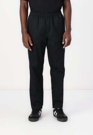 Black athletic pants with an elastic waistband, casual fit, two side pockets, and lightweight fabric are worn with black sneakers.