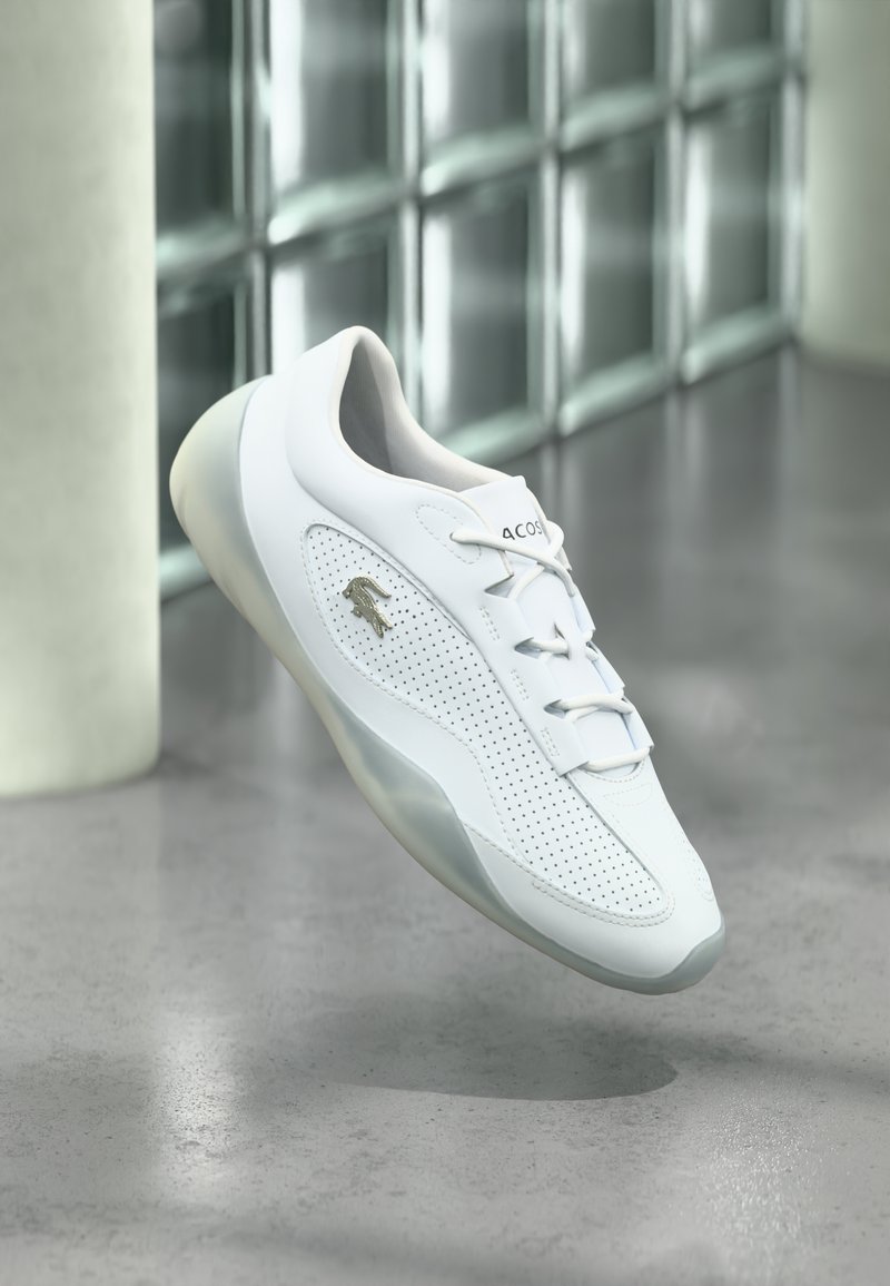 Baskets blanches Lacoste avec côtés perforés et logo crocodile argenté, flottant au-dessus d'un sol en béton poli près d'un mur en parpaings de verre.