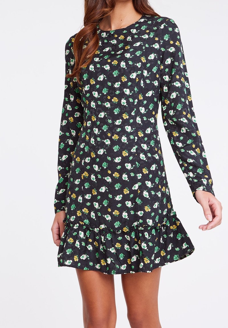 Femme portant une robe noire à manches longues avec des fleurs vertes, blanches et jaunes et un ourlet volanté, debout devant un fond blanc.