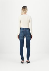 Woman with long blonde hair stands facing away wearing a cream ribbed sweater, dark blue skinny jeans, and black heeled sandals.