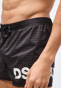 Primer plano de una persona con pantalones cortos de baño negros de DSQUARED2, con cinturilla elástica y cordón, mostrando el torso y la mano derecha.