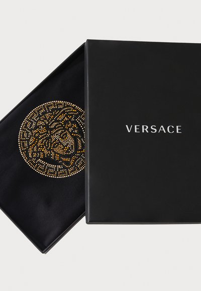 Fekete Versace doboz enyhén nyitva, fekete szövet belsővel, amely kör alakú arany és ezüst kristálymintát mutat. A márkanév fehérrel látható.