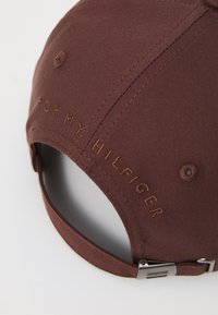 Brown cotton cap featuring embroidered "TOMMY HILFIGER" text, adjustable metal buckle, and a curved brim. Smooth texture and solid design.