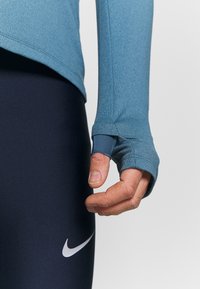 Top atletico blu a maniche lunghe con fori per i pollici e una finitura testurizzata, abbinato a leggings attillati blu navy con un logo bianco sul lato.