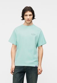 Νεαρός με σκούρα μαλλιά, φορώντας ανοιχτό μπλε μπλουζάκι με σχέδιο και σκούρο τζιν, στέκεται μπροστά σε λευκό, απλό φόντο.