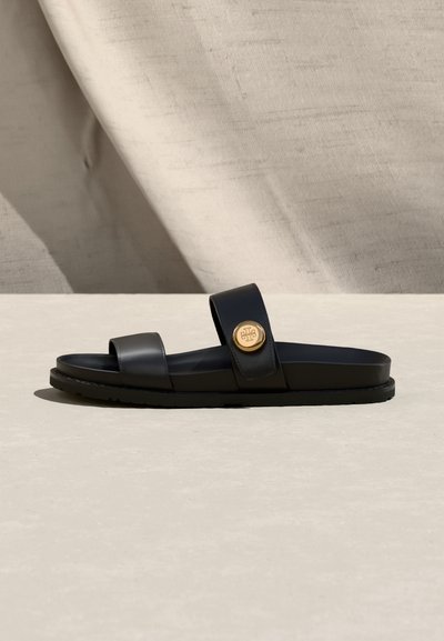Μαύρη επίπεδη σανδάλι με δύο λουριά, το ένα με στρογγυλό χρυσό έμβλημα, τοποθετημένη σε ανοιχτή ουδέτερη επιφάνεια και φόντο.
