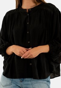 Blusa nera con bottoni, in un tessuto testurizzato, caratterizzata da un collo rotondo, dettagli plissettati e maniche a sbuffo. Bottoni dorati decorano la parte anteriore.