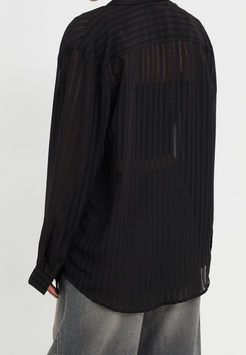 Personne portant une chemise noire transparente à manches longues avec des rayures verticales et un pantalon large gris, vue de dos sur un fond uni.