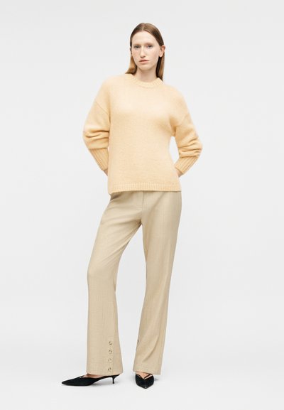 Femme debout portant un pull beige à poils, un pantalon beige clair avec des boutons près des chevilles, et des chaussures noires à talons bas pointues sur un fond blanc.