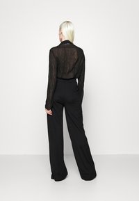 Zwarte transparante blouse met lange mouwen, gecombineerd met hoogwezige, wijduitlopende zwarte broek. Beide kledingstukken hebben een gladde textuur en een minimalistisch ontwerp.