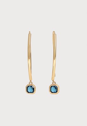 Longues boucles d'oreilles en or avec une petite pierre précieuse bleue carrée suspendue en bas sur un fond clair uni.