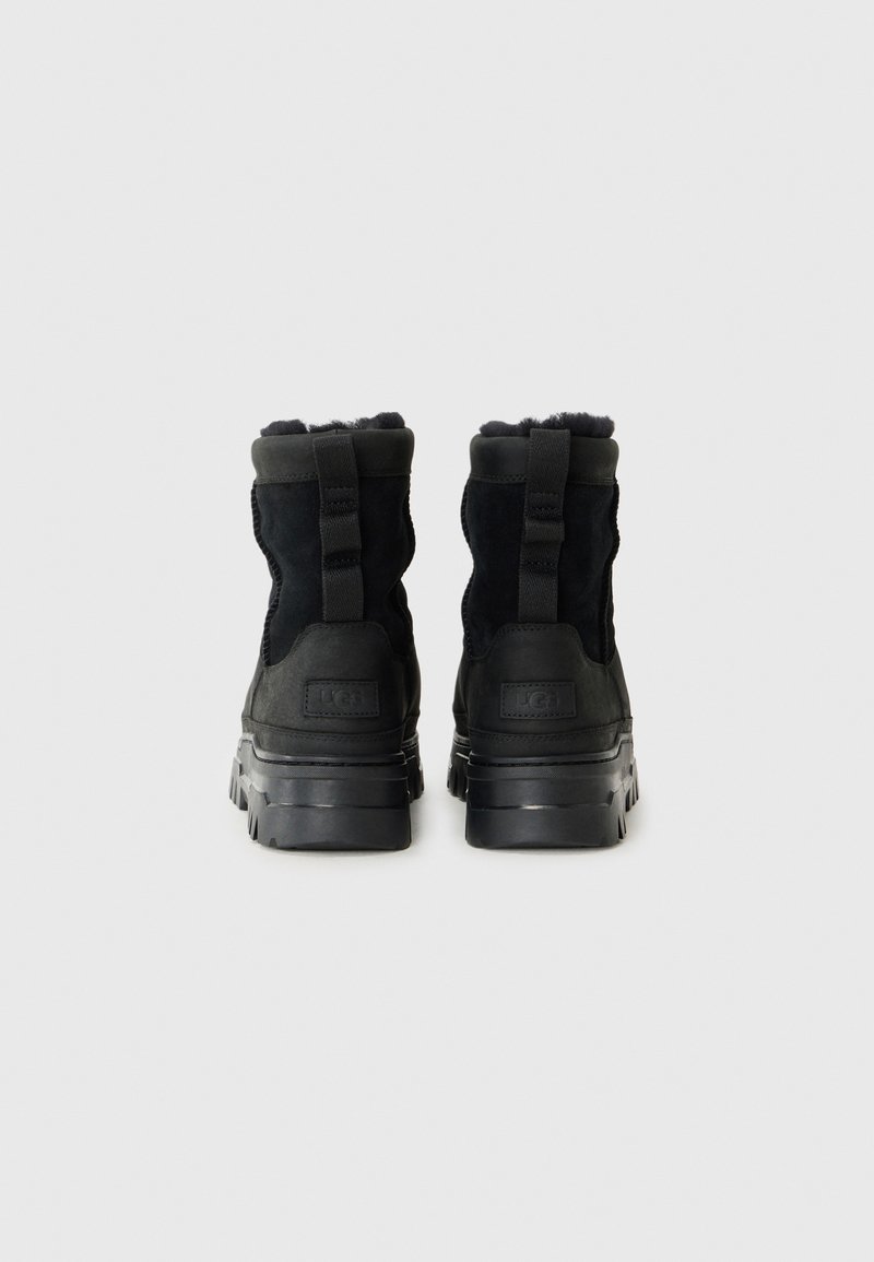 Schwarze Knöchelstiefel mit strukturiertem Obermaterial und klobigen Sohlen, ausgestattet mit Zuglaschen und weicher Innenauskleidung, von hinten gegen einen schlichten Hintergrund präsentiert.