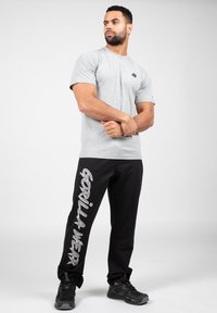 Grijze t-shirt met korte mouwen en een zwart logo, gecombineerd met zwarte joggingbroek met witte tekst "GORILLA WEAR" en zwarte sneakers.