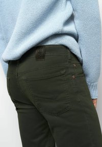 Person, der olivgrüne Jeans und einen hellblauen Strickpullover trägt, der die Gesäßtasche und den Bund mit einem Lederpatch zeigt.