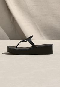 Μαύρο σανδάλι πλατφόρμας με σφήνα, λουράκι δαχτύλου και διακοσμητική κυκλική οπή πάνω σε ουδέτερο φόντο με υφασμάτινο παρασκήνιο.