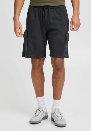 Pantaloni scurți negri dintr-un material moale, cu buzunare laterale, talie elastică și o croială lejeră. Purtați cu adidași gri și șosete albe.