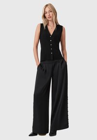 Kobieta stoi z rękami w kieszeniach, ubrana w czarną bezrękawnikową koszulę zapinaną na guziki, szerokie czarne spodnie oraz czarne czółenka na szpic, na szarym tle.