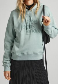 Felpa verde menta con cappuccio e logo frontale "Pepe Jeans London", caratterizzata da una vestibilità comoda, cappuccio con coulisse e polsini a costine, abbinata a una gonna nera plissettata.