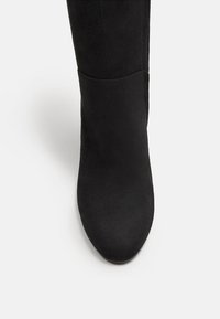 Botte en suède noire, haute jusqu'au genou, avec un bout arrondi et une texture lisse et mate. Design simple sans aucune quincaillerie ou embellissements visibles.
