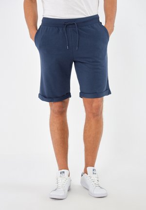 Uomo con pantaloncini blu navy con coulisse e sneakers bianche in piedi con le mani nelle tasche su uno sfondo semplice.