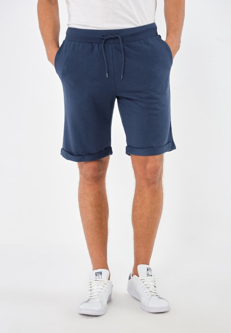 Homme portant un short bleu marine avec cordon et des baskets blanches, debout, mains dans les poches, sur un fond uni.