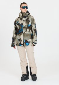 Skijacke mit mehrfarbigem Camouflage-Muster in Braun-, Blau- und Grautönen. Kombiniert mit hellbeigen Hosen und schwarzen Skischuhen.