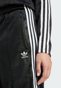 Zwarte Adidas trainingspak met witte drie-streepaccenten en logo. Voorzien van een ritszak aan de zijkant en een zachte, gladde textuur.