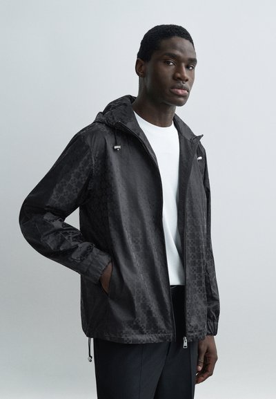Homme portant une veste à capuche noire avec un motif quadrillé subtil sur une chemise blanche, debout avec une main dans la poche devant un fond uni.