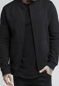 Felpa nera con una texture liscia, chiusura con zip frontale, polsini a costine, tasche laterali e un logo ricamato sul lato sinistro del petto.