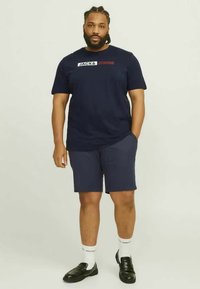 Camiseta de algodão azul-escuro com logótipo branco e vermelho, combinada com calções azul-escuro. Sapatos mocassins pretos e meias brancas com detalhe do logótipo completam o look.