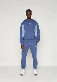 Mėlynas džemperis su šviesiai mėlynais šonų akcentais ir logotipu; derančios sportinės kelnės su panašiais akcentais; medžiaga atrodo minkšta; modelis dėvi baltus sportinius batelius.