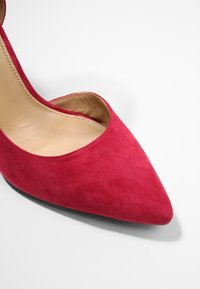 Scarpa con tacchi alti in suede rosso, con punta affilata e design a scollatura bassa, caratterizzata da un interno liscio e un esterno testurizzato.
