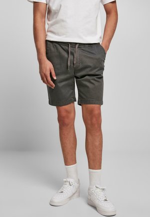 Homme portant un short gris foncé avec cordon, des chaussettes blanches hautes, des baskets blanches et un t-shirt blanc à manches courtes, debout avec une main dans la poche.