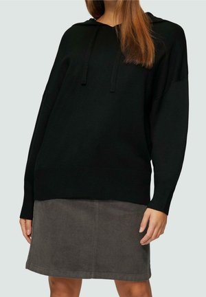 Kobieta w czarnej, długorękawowej bluzie z kapturem i szarej spódnicy sztruksowej do kolan, stojąca na tle gładkiego, jasnego tła.