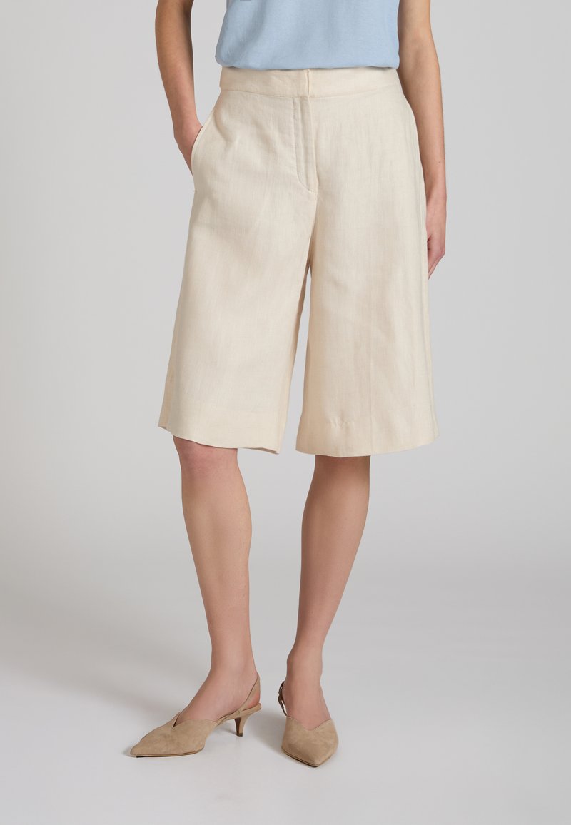 Mujer vistiendo pantalones cortos beige de pierna ancha hasta la rodilla, camiseta azul claro y tacones beige puntiagudos con tira trasera, de pie con una mano en el bolsillo.
