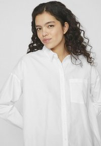 Camisa blanca de botones con mangas largas, bolsillo frontal y textura suave, que presenta un cuello clásico y un ajuste holgado y relajado.