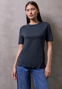 Giovane donna con lunghi capelli marroni che indossa una maglietta a maniche corte blu scuro e jeans blu, in piedi contro un muro di cemento grigio chiaro.
