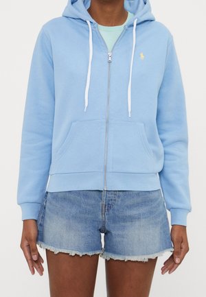 Persoon draagt een lichtblauwe hoodie met rits en witte trekkoorden, gecombineerd met spijkershort met rafelige zoom, en staat met de armen ontspannen langs het lichaam.