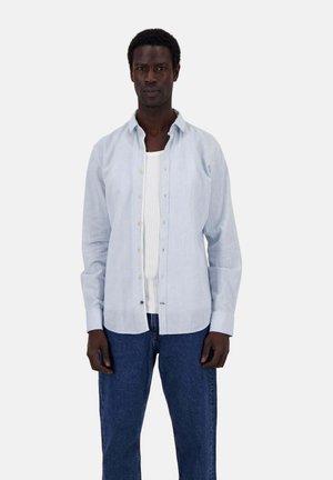 Helles blaues Langarmhemd mit durchgehender Knopfleiste, aus einem weichen, strukturierten Stoff; getragen über einem weißen Tanktop und kombiniert mit dunklen Jeans.