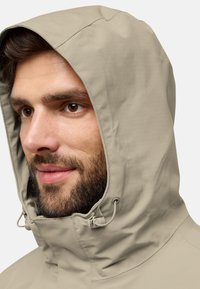 Giacca beige con cappuccio e superficie texturizzata, dotata di cordini regolabili al collo e una vestibilità aderente attorno al viso.