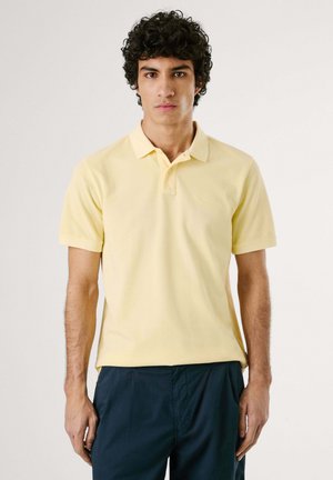 Kollane puuvillane polo särk lühikeste varrukatega, kraega ja diskreetse logoga rinnaosal, koos tumedate mereväe värvi pükstega.