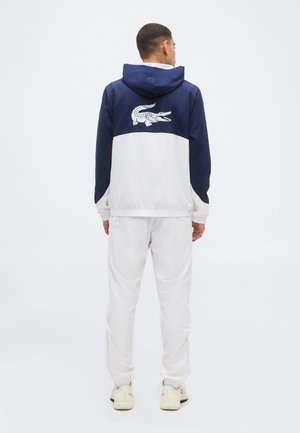 Homme portant une veste à capuche bleu marine et blanche avec un grand logo crocodile dans le dos, pantalon blanc et baskets crème, debout de dos.