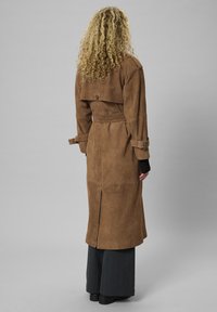 Göndör szőke hajú személy hosszú barna velúr trench coatban, fekete kesztyűben, sötét nadrágban és fekete cipőben, háttal állva.