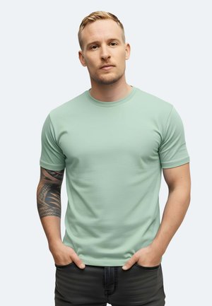 Mintgrünes T-Shirt mit kurzen Ärmeln und rundem Halsausschnitt, aus weichem Material. Das Model hat ein Tattoo am rechten Arm und trägt dunkle Hosen.