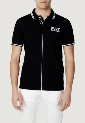 Homme portant un polo noir à manches courtes avec une bordure blanche et le logo EA7 Emporio Armani, associé à un pantalon blanc, debout devant un fond uni.