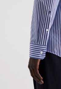 Braccio destro di una persona che indossa una camicia a righe verticali blu e bianche e pantaloni scuri, mano rilassata al suo fianco.